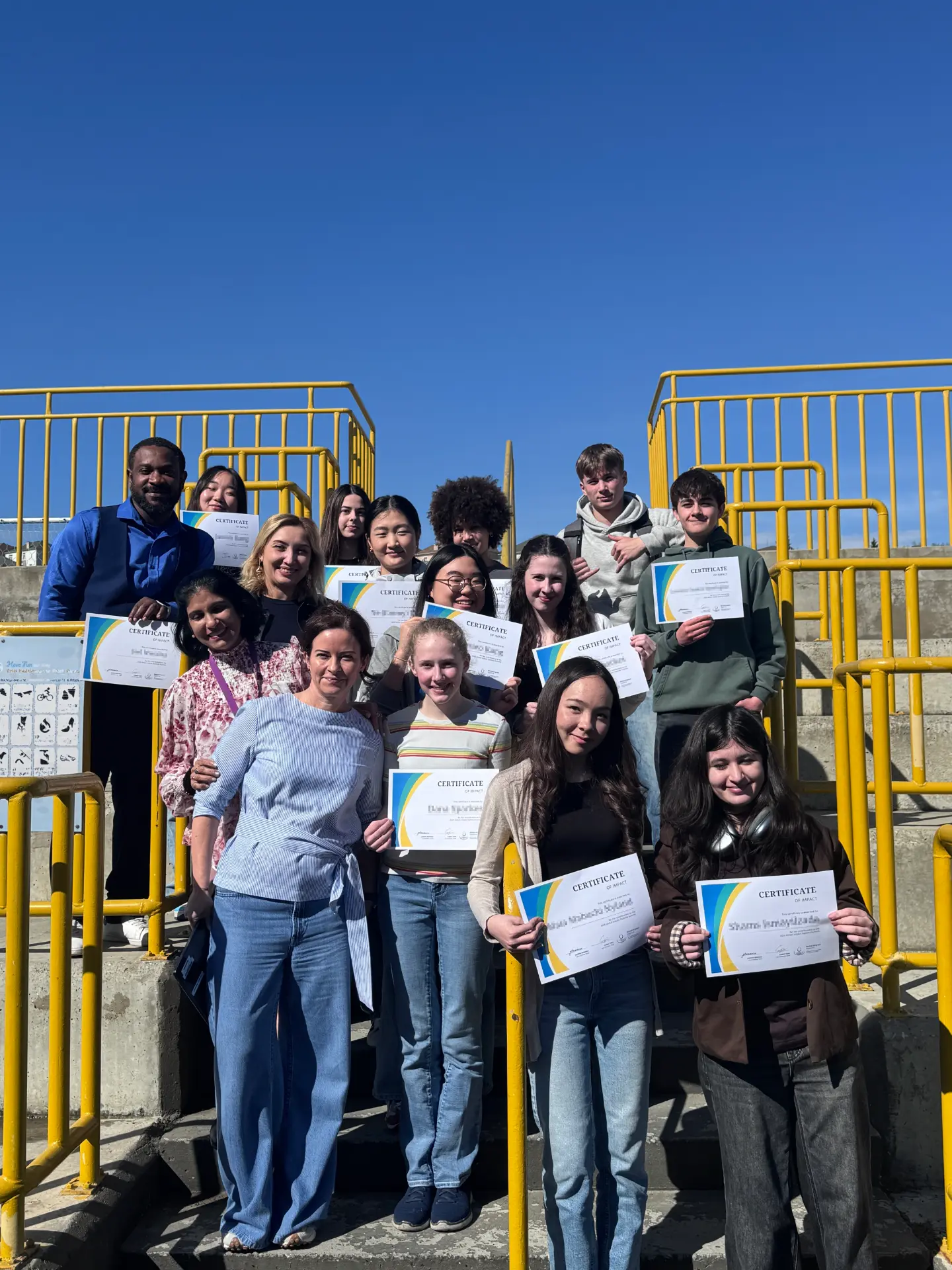 GID Student Leaders, including Rebecca Greenall, pose on an outdoor staircase holding their Certificate of Impact awards against a clear blue sky.