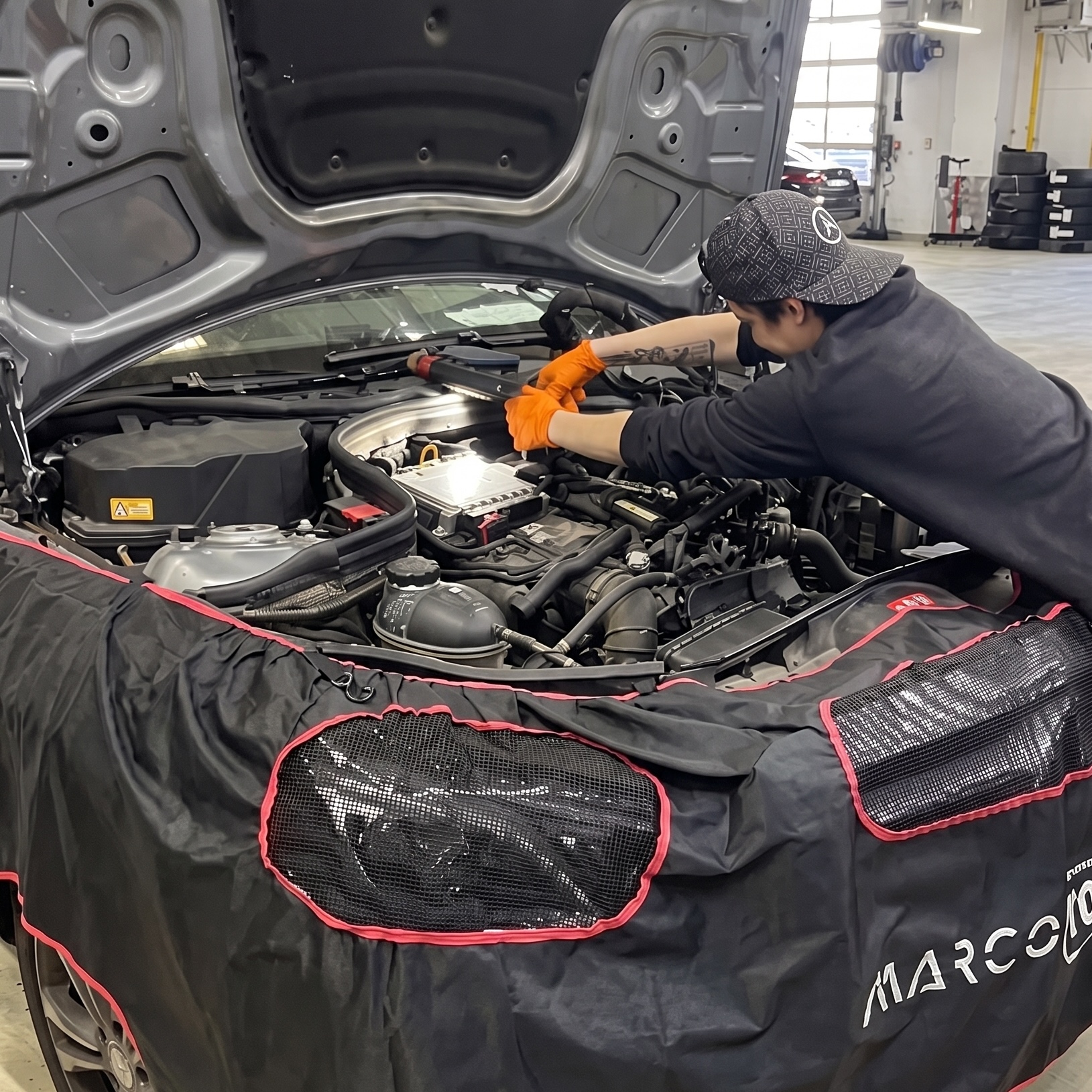A student from AIS Bucharest works on a car engine during her internship, wearing orange work gloves in a professional garage.