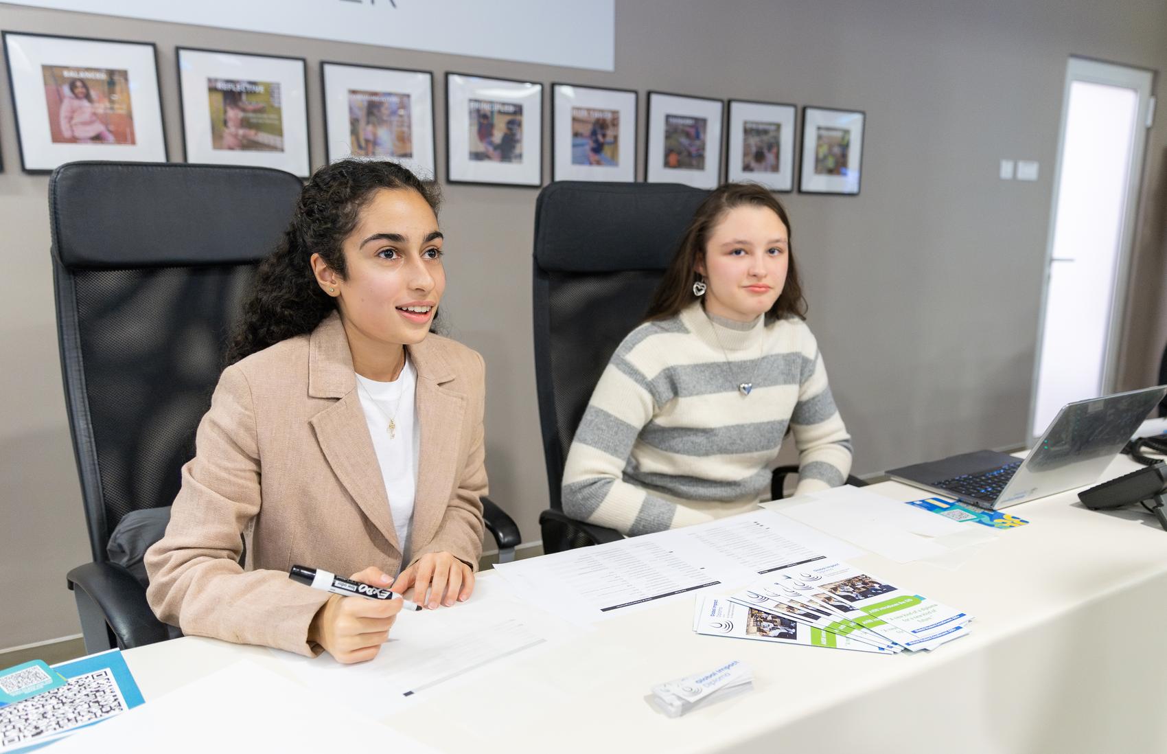 Two students sit behind a table of Global Impact Diploma leaflets at a school event, one speaking to visitors while the other listens.